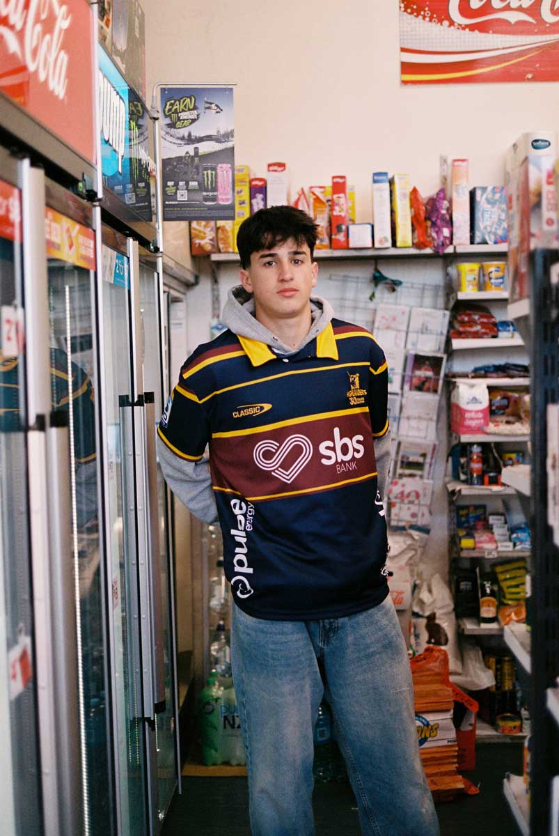 A boy in a dairy next to fridges wearing a highlanders jersey